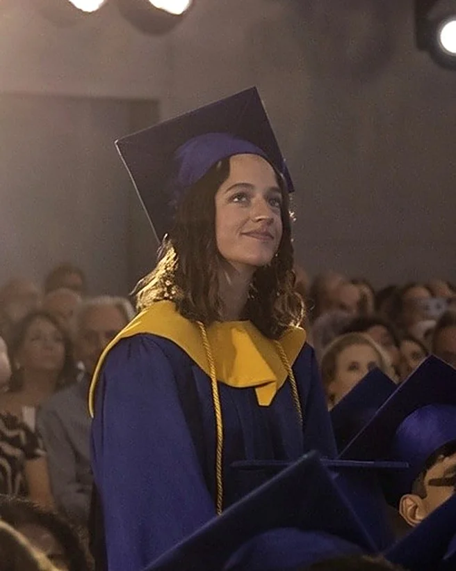 Marian Garabana standing among seated graduates at her graduation ceremony, wearing a blue cap and gown with a yellow stole